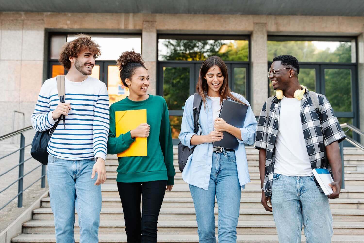 Four diverse college students walking down stairs outside a campus building, smiling and holding folders, books, and backpacks, enjoying a lively conversation.