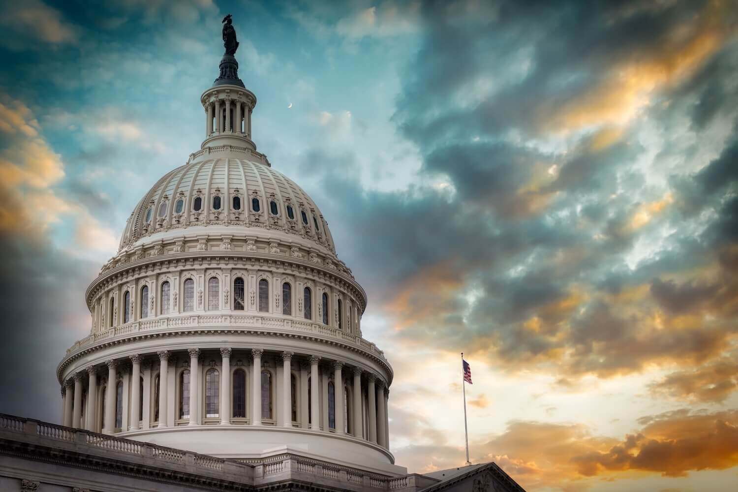 The United States Capitol dome under a dramatic sunset sky with clouds, featuring the Statue of Freedom on top and an American flag to the side.